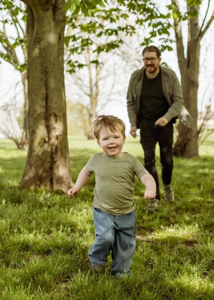 A laughing toddler with strawberry blonde hair runs across a sunlit lawn in an olive t-shirt and blue trousers, with his father in a green shirt and glasses following behind through the trees.