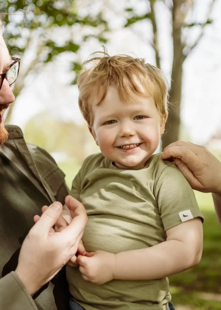 A close-up of a smiling toddler with strawberry blonde hair, wearing an olive t-shirt, held in the arms of his father whose green-shirted shoulder and bearded profile frame one side of the shot.