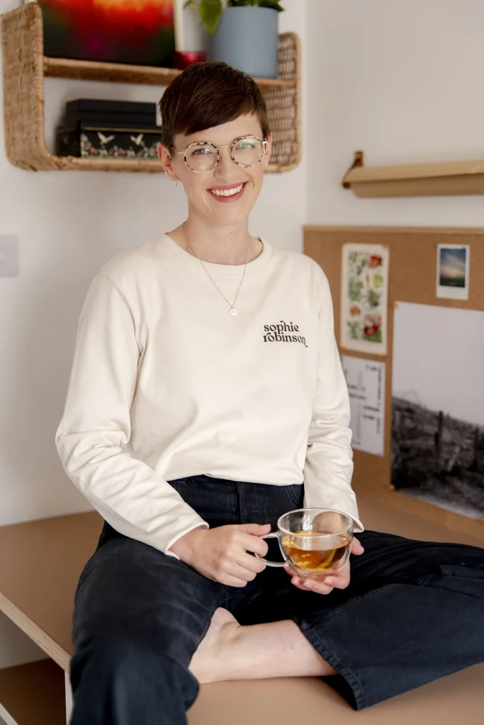 A woman with a short brown pixie cut and tortoiseshell glasses sits cross-legged on a desk, smiling at the camera and holding a glass mug of tea with a lemon slice. She wears a cream long-sleeved top branded "sophie robinson" and dark jeans, with a rattan shelf and pinboard of artwork behind her.