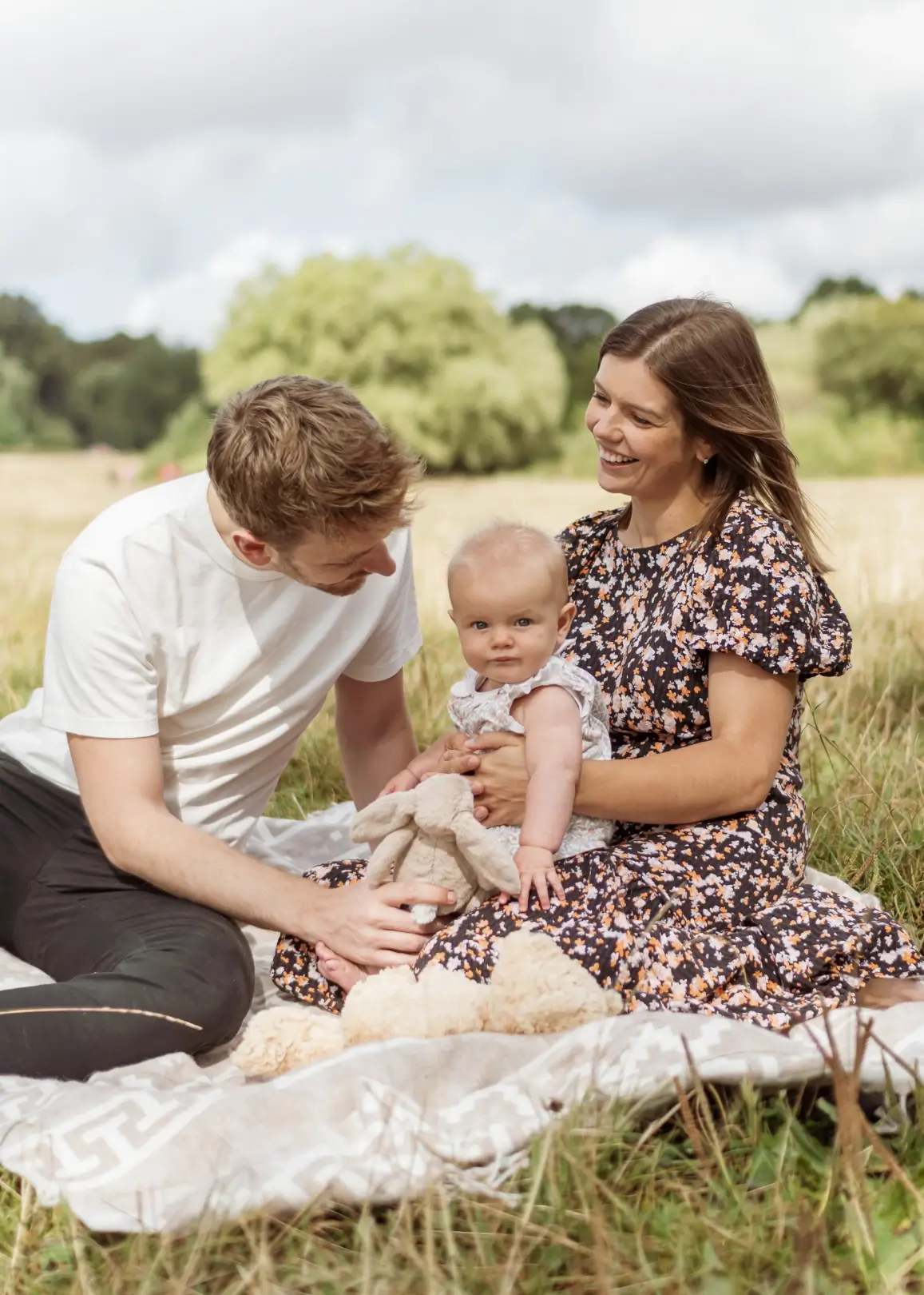 A young couple sit on a picnic blanket in a meadow with their baby daughter, the mother in a floral dress holding her in her lap while the father in a white t-shirt leans in smiling. A plush bunny rests beside them on the blanket.