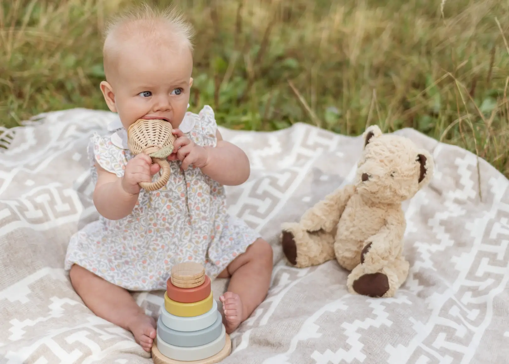 Close-up of a baby girl in a floral dress sitting on a cream patterned blanket, chewing on a wooden rattle teether. A wooden stacking toy and a teddy bear sit beside her.