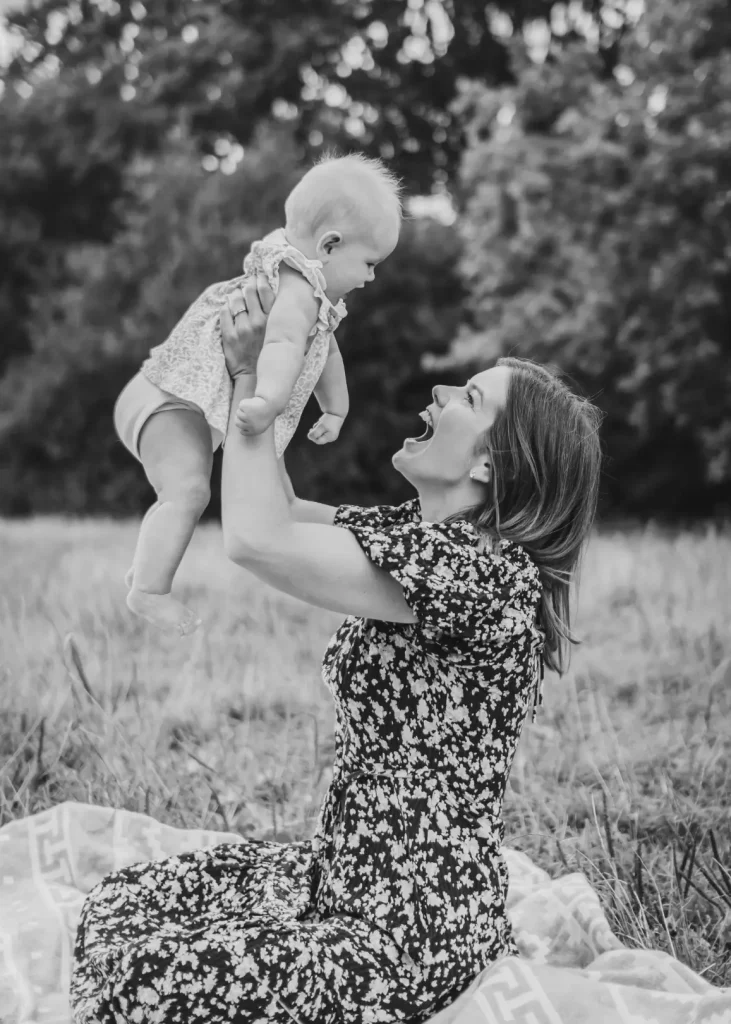 Black and white photograph of a mother in a floral dress kneeling on a picnic blanket in a meadow, lifting her laughing baby daughter high into the air as they grin at each other.
