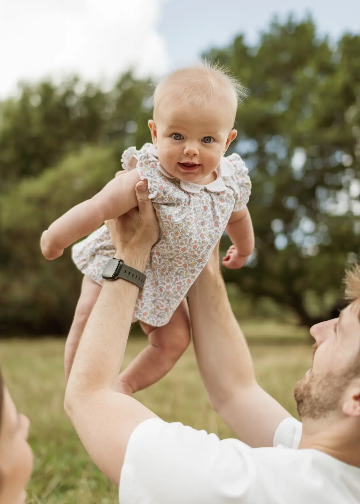 A father in a white t-shirt lifts his baby daughter up into the air, the baby grinning down at him as she's held aloft against a backdrop of green trees and soft sky.
