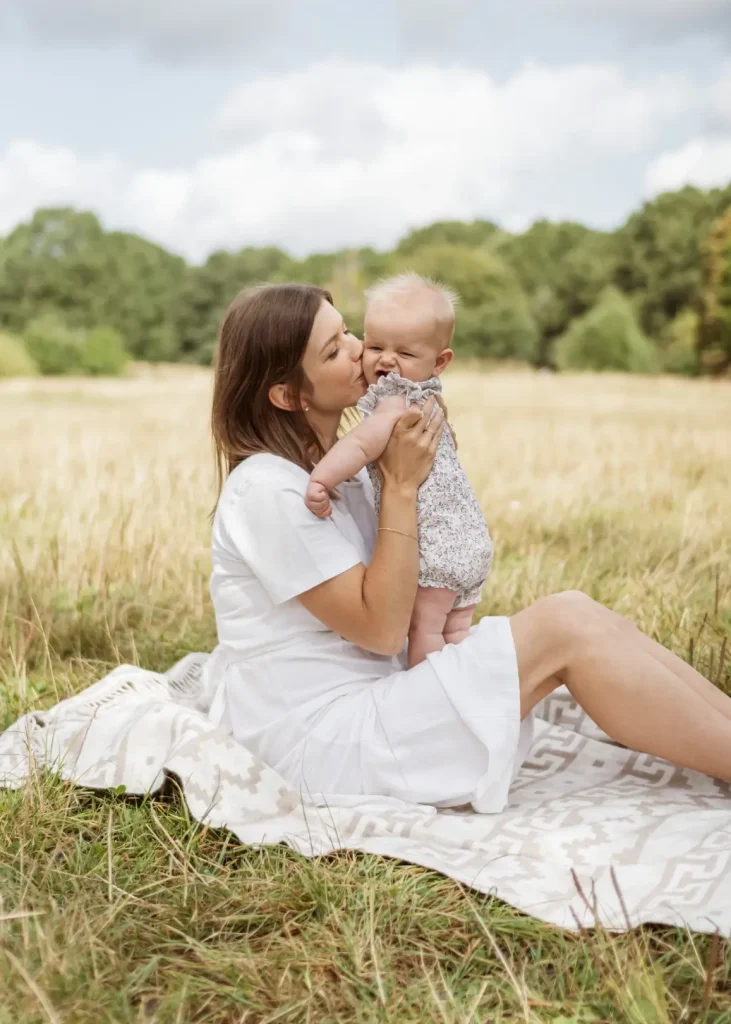 A mother in a white dress sits on a picnic blanket in a meadow, kissing her giggling baby daughter on the cheek as she holds her up. Long golden grasses fill the background.