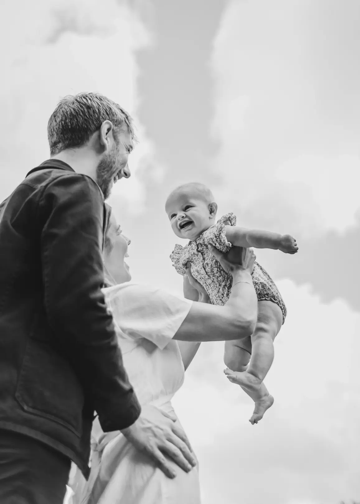 Black and white photograph taken from a low angle, looking up at a couple lifting their laughing baby daughter into the air against a cloudy sky, both parents smiling up at her with delight.