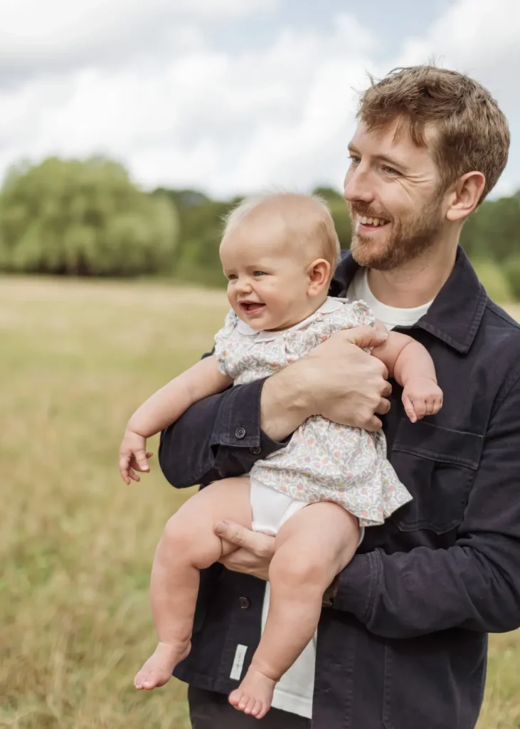 A father with a beard, wearing a dark jacket over a white t-shirt, holds his laughing baby daughter on his hip in a sunlit meadow, both looking off to one side.
