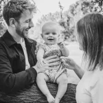 Black and white photograph of a couple standing on either side of their baby daughter, who is perched on a sturdy tree branch in a floral romper, giggling at the camera as her parents smile at her.