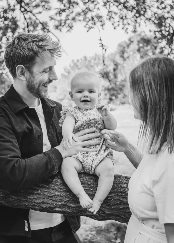 Black and white photograph of a couple standing on either side of their baby daughter, who is perched on a sturdy tree branch in a floral romper, giggling at the camera as her parents smile at her.