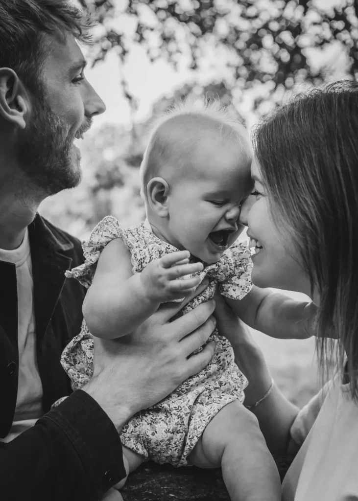 Black and white close-up of a couple sharing a joyful moment with their baby daughter, the mother pressing her forehead to the baby's cheek as the baby laughs with her mouth wide open.