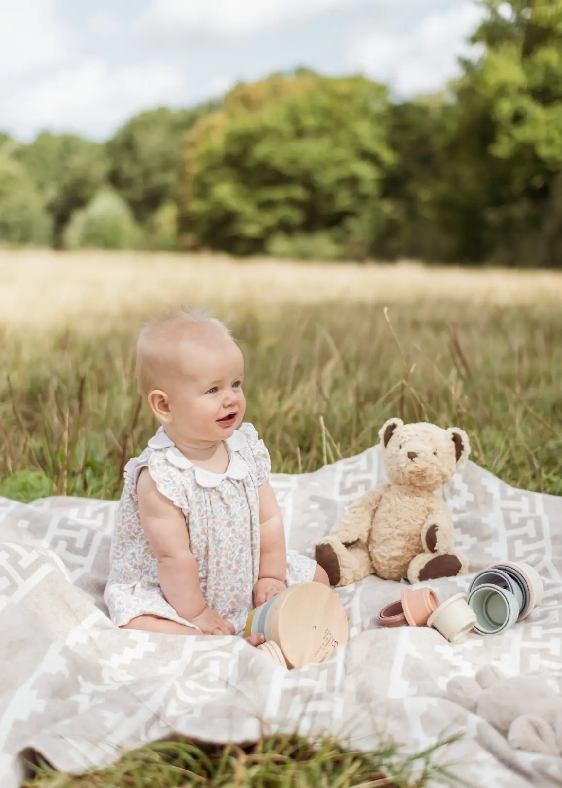 A baby girl in a floral dress with a white collar sits on a cream patterned blanket in a meadow, smiling and looking off to one side. A teddy bear and a tumbled wooden stacking toy lie beside her.