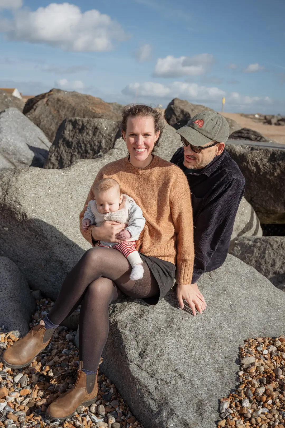 A couple sit together on a large grey boulder on a pebbled beach, the mother in a rust-coloured jumper holding their baby on her lap, the father beside her in a green cap and dark corduroy shirt. The sea is just visible in the background.