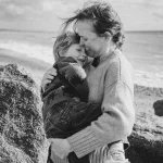 Black and white photograph of a mother in a knitted jumper hugging her young son close on a pebbled beach, both laughing as they press their foreheads together with the sea behind them.