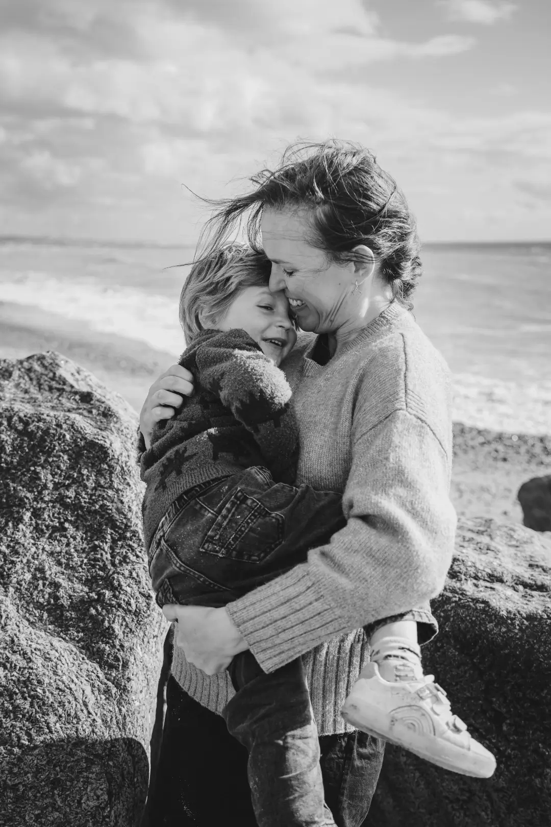 Black and white photograph of a mother in a knitted jumper hugging her young son close on a pebbled beach, both laughing as they press their foreheads together with the sea behind them.
