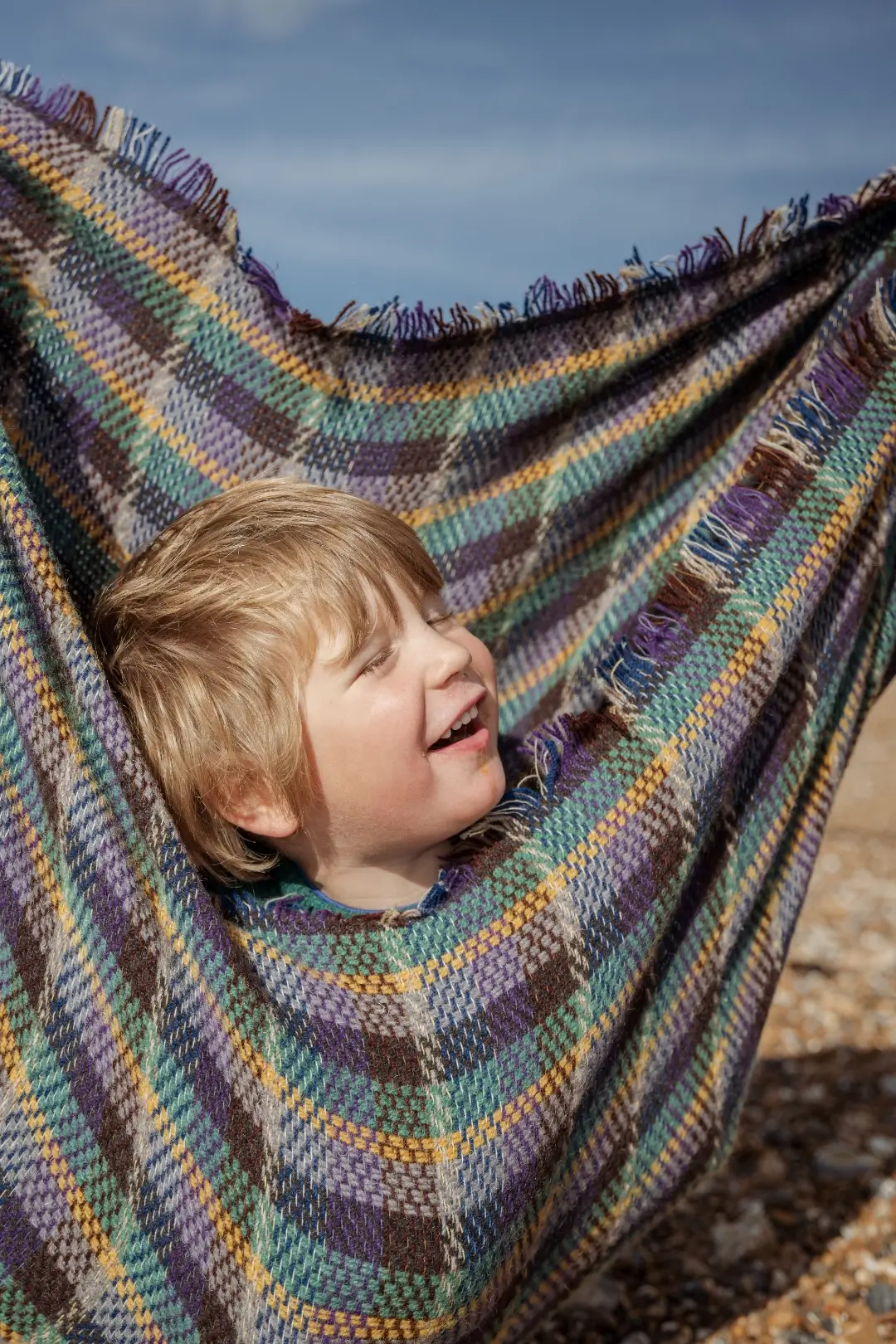 A blonde young boy laughs as he's cradled in a colourful checked wool blanket held up like a hammock, with a blue sky and pebbled beach behind him.