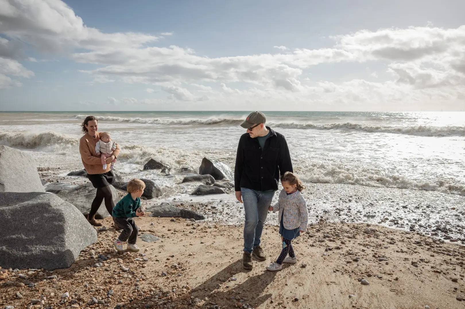 A young family walks along a pebbled beach with crashing waves behind them. The mother in a rust-coloured jumper carries the baby, while the father in a dark shirt and cap walks hand-in-hand with their young daughter, and their son explores the rocks nearby.
