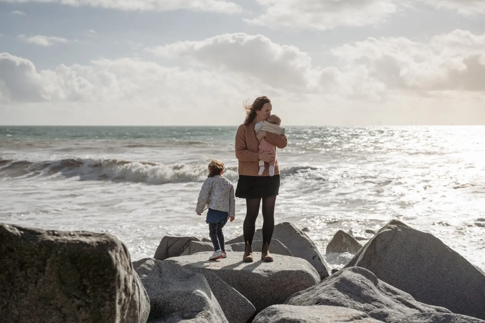 A mother in a rust-coloured jumper stands on the sea-defence boulders kissing her baby on the head, while her young daughter stands beside her looking out at the breaking waves.