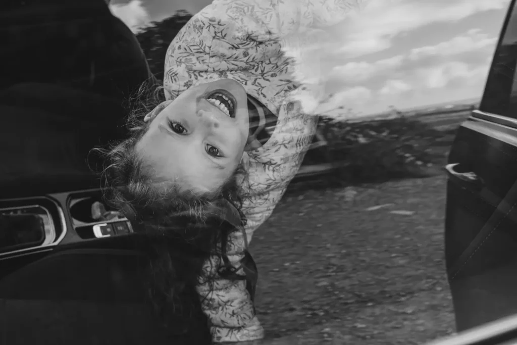 lack and white photograph of a young girl hanging upside down out of the open door of a car, laughing with her hair tumbling toward the ground.