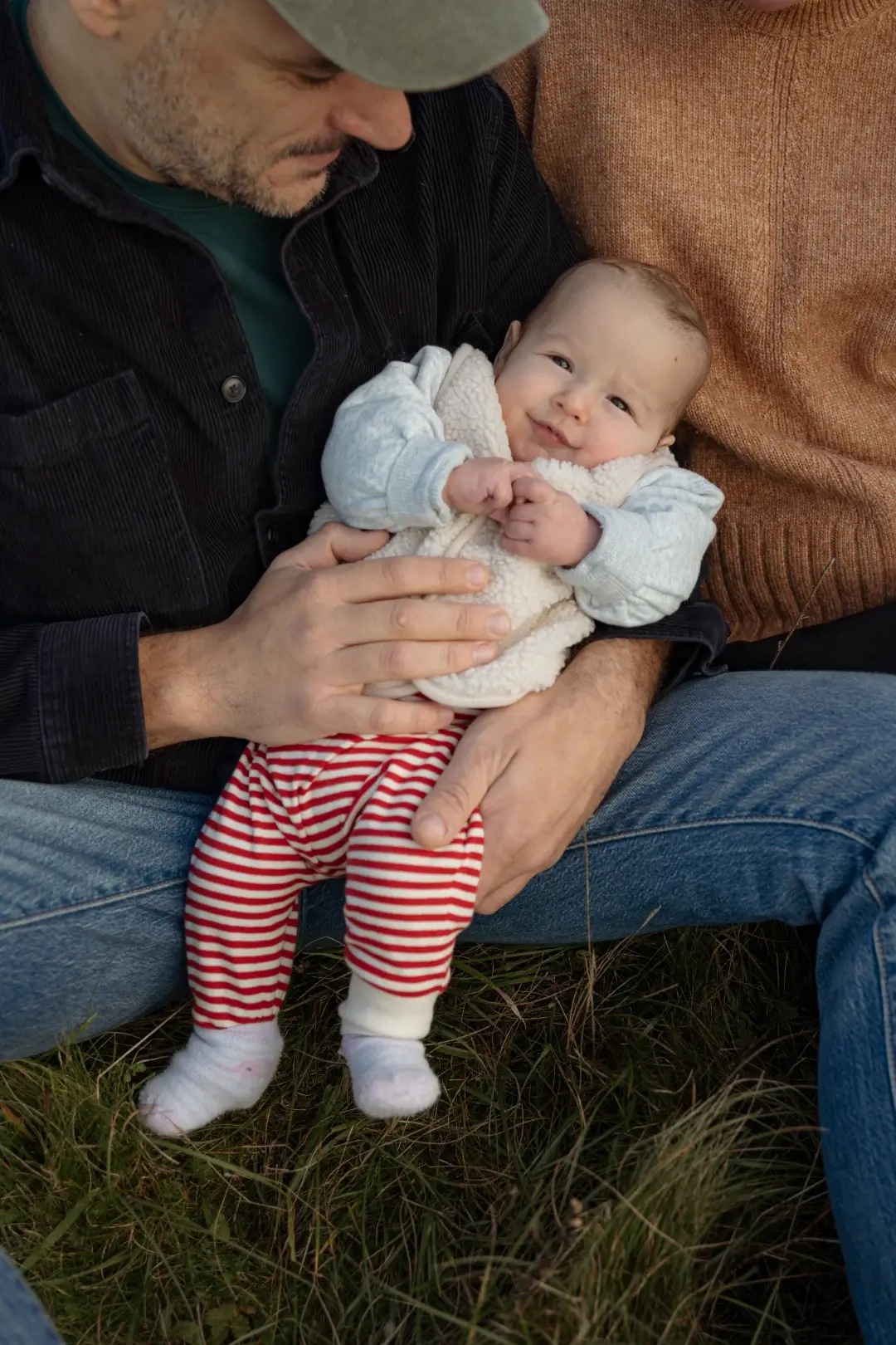 A father in a green cap and dark corduroy shirt looks down lovingly at his baby, who stands supported on his lap wearing a cream fleece jacket and red striped trousers.