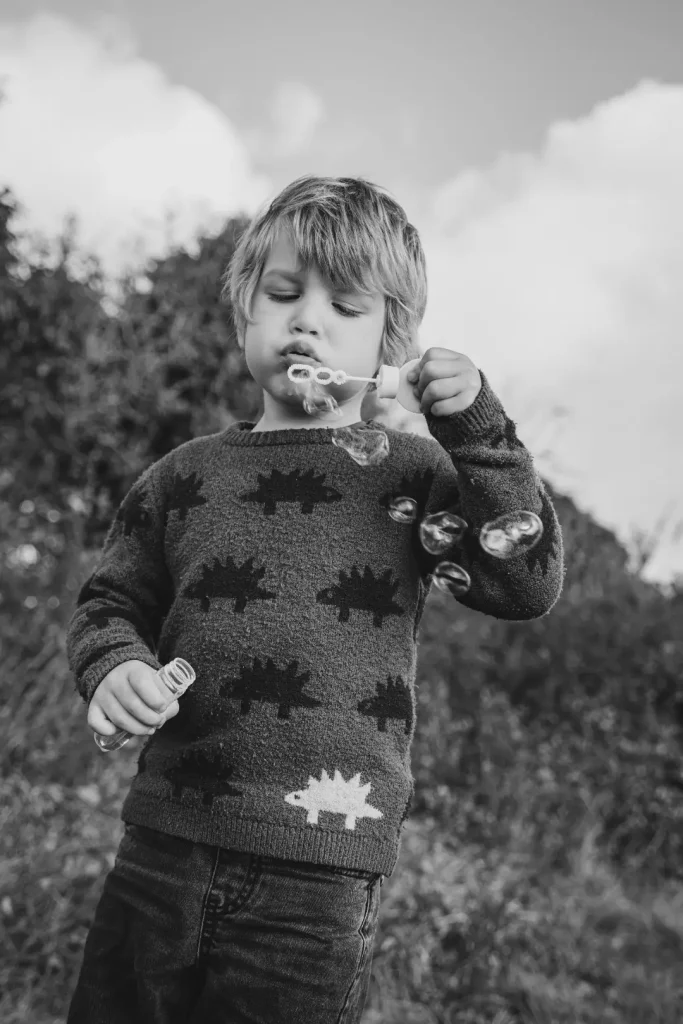 Black and white photograph of a young boy in a dinosaur-patterned jumper blowing bubbles, concentrating on the wand as the bubbles drift around him.