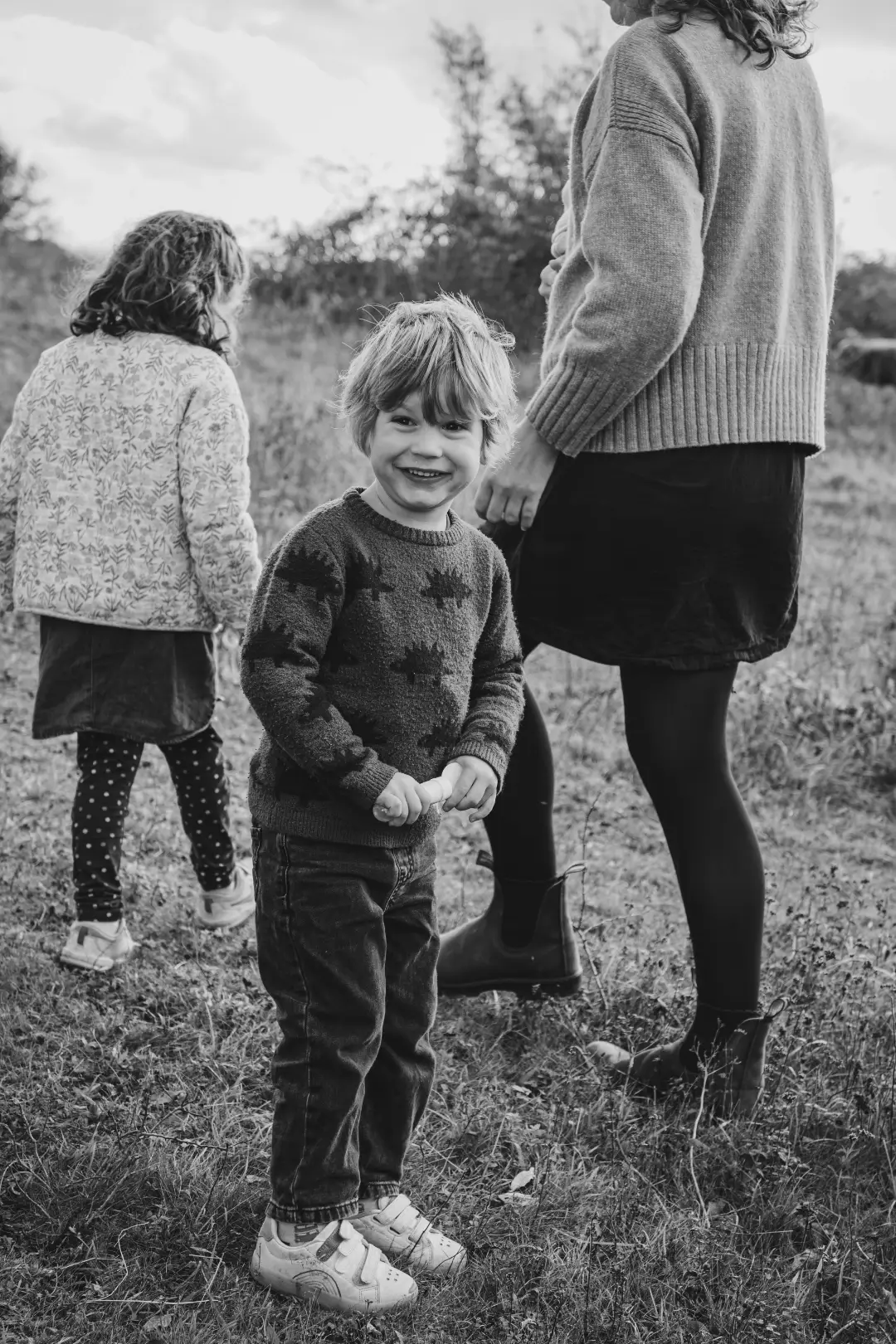 Black and white photograph of a young blonde boy in a dinosaur-patterned jumper grinning at the camera, holding his mother's hand, while his sister wanders just behind in a floral jacket.
