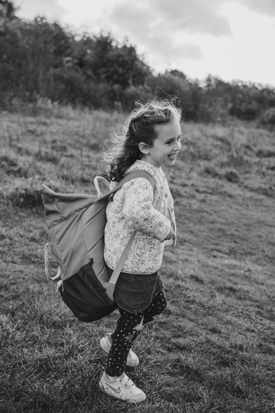 Black and white photograph of a young girl with long hair running through a grassy field, laughing, with a large backpack over her shoulders.