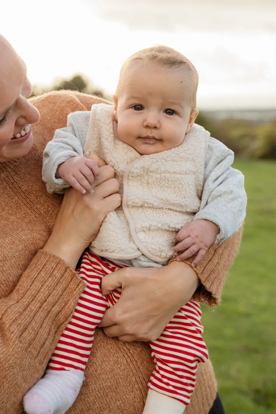 A baby wearing a fleece gilet over a grey top and red striped trousers is held close by her smiling mother in a rust-coloured jumper, against a soft outdoor backdrop of greenery and sky.