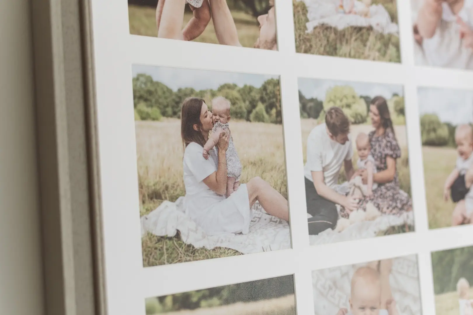 A close-up of a multi-image framed print mounted on a wall, showing several photographs from an outdoor family session. The most prominent image shows a woman with long dark hair wearing a white dress, sitting on a white blanket in a meadow and kissing a baby she holds in her arms. To the right, a partially visible photograph shows a man in a white t-shirt and a woman in a floral dress sitting together on a blanket with the same baby. Partial views of further images are visible at the edges of the frame.