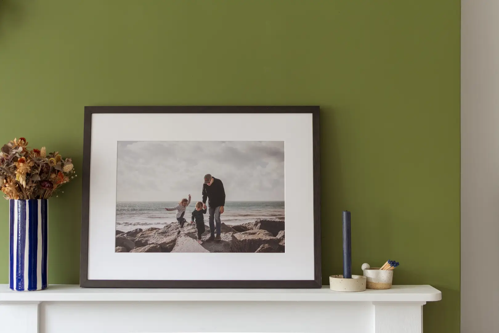 A large dark-framed print with a white mount leans against an olive green wall on a white mantelpiece. The photograph inside shows an adult and two young children playing on a rocky shoreline beneath a cloudy sky, with the sea visible behind them. To the left of the frame sits a blue and white striped ceramic vase holding dried flowers in amber, rust, and cream tones. To the right, a slim navy pillar candle stands beside two small ceramic pots, one holding matchsticks.