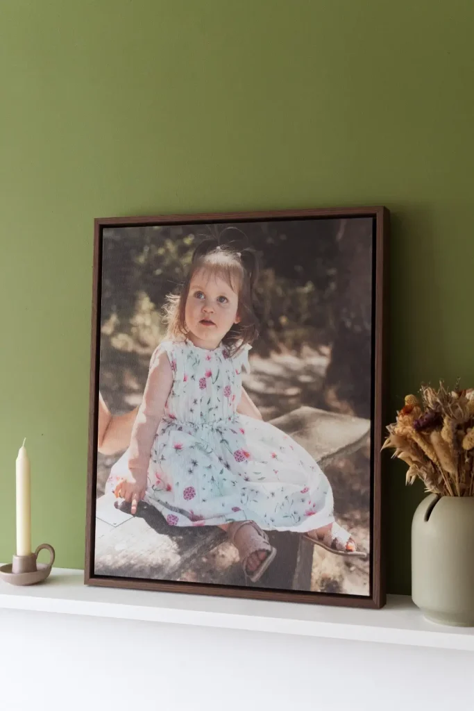 A large canvas print in a dark walnut frame leans against an olive green wall on a white shelf. The canvas shows a portrait of a toddler girl with light brown hair in a small ponytail, wearing a white floral sundress with pink and purple flower details and tan sandals. She sits outdoors in dappled sunlight, looking upwards with wide blue eyes and a curious expression. To the left of the canvas, a cream pillar candle sits in a small clay candle holder. To the right, a sage green ceramic vase holds a bunch of dried flowers and grasses in warm amber and dusty pink tones.
