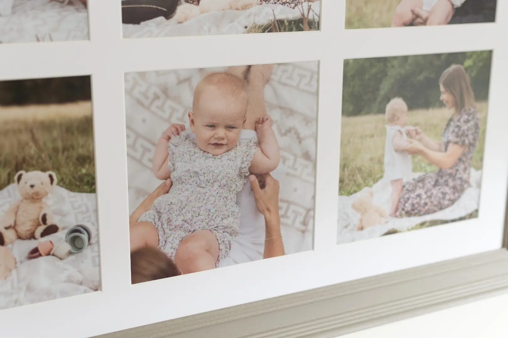 A close-up of a multi-aperture grey wooden frame showing three of its photographs. The central and most prominent image shows a baby in a pale floral romper suit being held up by an adult's hands, smiling contentedly at the camera. To the left, a partial image shows a cream teddy bear and a small cup on a white patterned blanket in a meadow. To the right, a woman in a floral dress kneels on a blanket in a sunny field, holding the same baby up in front of her. The grey moulded frame edge is visible along the bottom of the image.