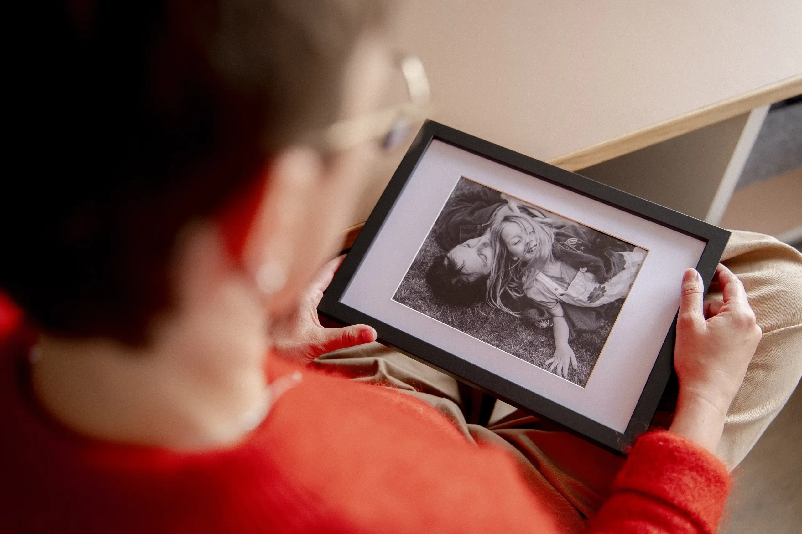A person with short dark hair and round tortoiseshell-framed glasses holds a black-framed, white-mounted print, looking down at it, viewed from over their shoulder. The print contains a black and white photograph of two people lying on grass, their heads close together, looking at each other. A warm beige desk surface and shelving unit are softly visible in the background.