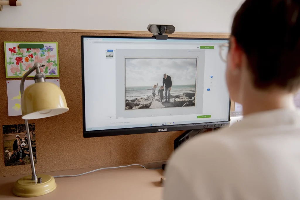 A woman with short dark hair and round tortoiseshell-framed glasses sits at a warm beige desk, viewed from behind over her shoulder, looking at an Asus monitor. The screen displays a photo editing application with a black and white photograph of an adult and two young children on a rocky beach. A yellow-green dome desk lamp, children's artwork, and family photographs are pinned to a corkboard behind the monitor. The person wears a cream long-sleeved top.