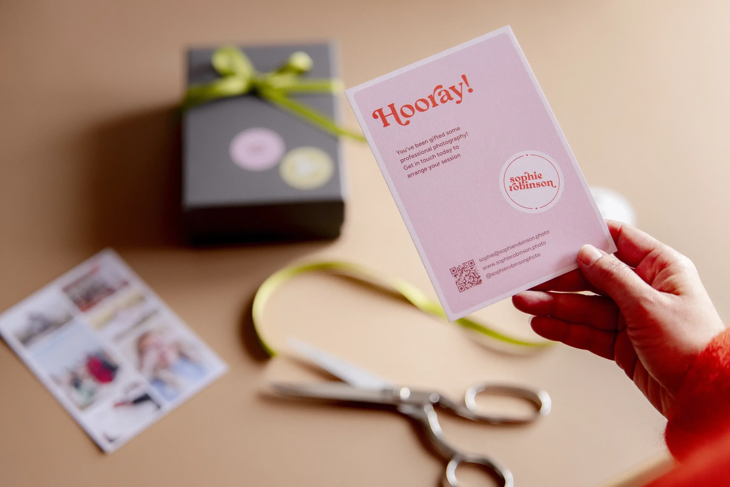 A hand with a red fluffy jumper sleeve holds up a blush pink gift card with "Hooray!" printed in red lettering at the top. The card reads "You've been gifted some professional photography! Get in touch today to arrange your session" and features the Sophie Robinson photography logo with contact details and a QR code. In the background, a dark grey gift box tied with a lime green ribbon sits on a tan surface, alongside a small printed photo card, a length of lime green ribbon, and a pair of silver scissors.