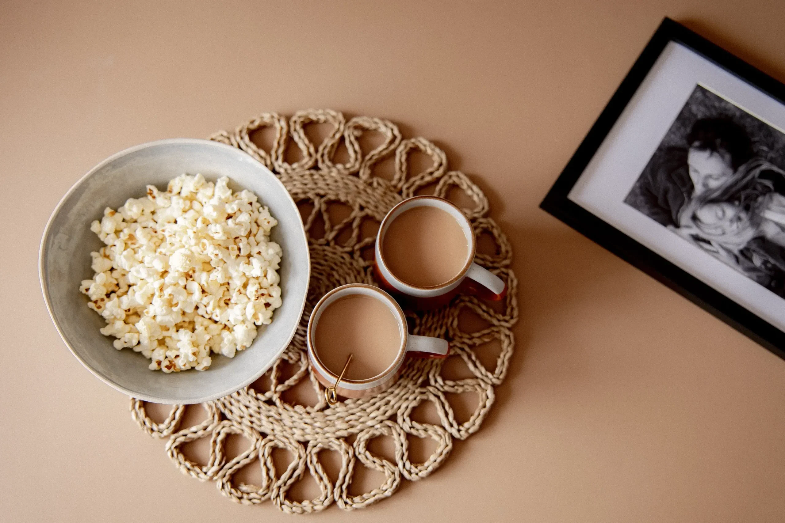 An overhead flat-lay on a tan surface showing two terracotta-rimmed mugs filled with milky tea and a grey ceramic bowl of popcorn, all arranged on a circular woven rattan placemat. To the left, a black-framed print displays a black and white photograph of two people in a tender embrace.