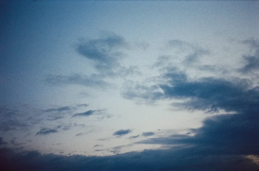 A 35mm film photograph of a dusk sky, shot with visible grain. Layers of dark blue-grey cloud drift across a pale blue-grey sky, with a brighter patch of light breaking through the cloud cover near the centre. The image has the characteristic colour shift and texture of film photography.