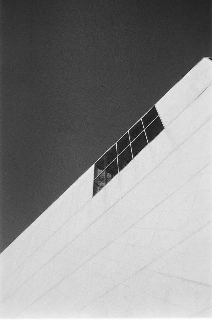 A black and white 35mm film photograph of the upper corner of a large angular building shot from below. The building's pale stone or concrete facade rises diagonally across the frame, with a band of gridded glazing set into the surface near the roofline. The sky above is a deep, even grey. The strong geometric composition and high contrast give the image a graphic, abstract quality. The image has a fine grain texture throughout.
