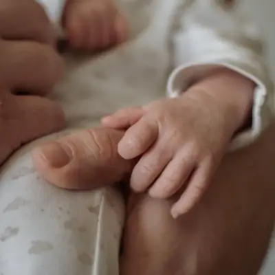 A close-up of a newborn baby's hand and foot resting together, with a parent's hand gently visible in the background. The baby wears a white outfit with a subtle pattern. The image is softly focused with a warm, natural tone.