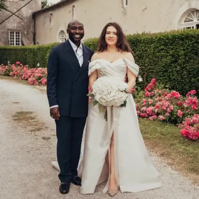 A couple stand together on a gravel path outside a stone country house, smiling at the camera. The man wears a navy double-breasted suit with a light tie, and the woman wears an off-the-shoulder ivory satin wedding dress with a front split, holding a large bouquet of white hydrangeas. Pink rose bushes in full bloom line the path behind them, with a neatly trimmed dark green hedge beyond.