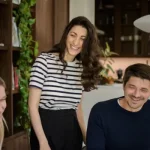 A woman in a striped top standing and laughing between a seated blonde woman and a man in a navy jumper. Trailing houseplants and wooden cabinetry frame the scene.