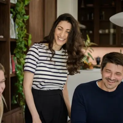 A woman in a striped top standing and laughing between a seated blonde woman and a man in a navy jumper. Trailing houseplants and wooden cabinetry frame the scene.
