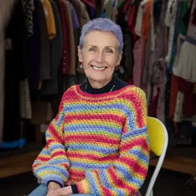 A woman with short lavender hair sits on a white folding chair, smiling warmly at the camera. She wears a brightly striped multicolour crochet jumper in red, yellow, blue, and green, and a black roll-neck underneath, with a beaded necklace. Behind her, rows of clothing on rails are visible in soft focus, suggesting a charity shop or vintage clothing setting.