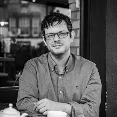 A black and white portrait of a man with dark hair and rectangular-framed glasses, seated at a table in a café or restaurant. He wears a grey button-up shirt and has his arms loosely folded on the table, with a teapot and cup visible in front of him. He looks directly at the camera with a relaxed expression. The background shows soft-focus café furniture and a window.