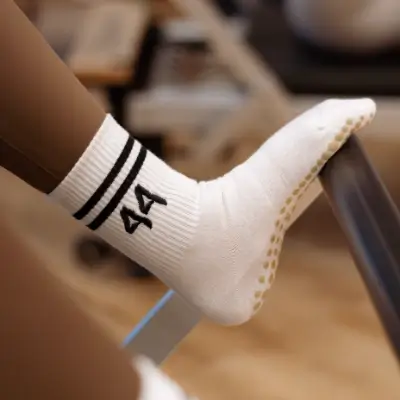 A close-up of a foot resting on a bar or ledge, wearing a white ribbed crew sock with two black stripes and the number 44 embroidered in black on the ankle. The sole of the sock has small yellow grip dots. The background is softly blurred, suggesting a fitness studio environment.